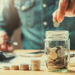 Businessman holding a coin, putting it into a glass jar, with his right hand holding a pencil and finger touching a calculator