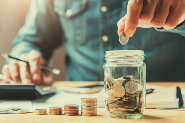 Businessman holding a coin, putting it into a glass jar, with his right hand holding a pencil and finger touching a calculator
