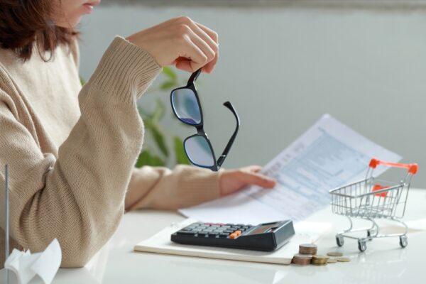 A woman sits at a table with her glasses in one hand looking at a printed copy of her bank statement with a calculator and coins next to it to understand her living costs